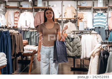 Young woman with long brown hair holds shopping bag while standing inside a clothing store, racks of apparel and accessories visible in the background 142452893