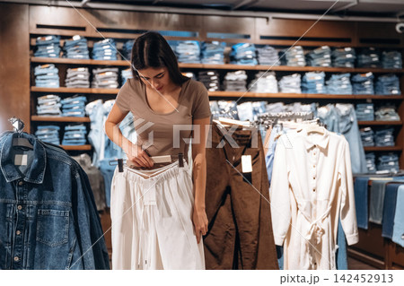 Female shopper examining white pants while standing in a clothing store with shelves of folded denim and garments in the background Female shopper examining white pants while standing in a clothing store with shelves of folded denim and garments in the background 142452913
