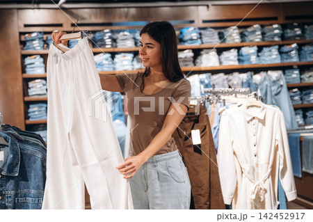 Young woman examining white pants while shopping in a clothing store, denim garments displayed on shelves in the background, casual fashion retail environment 142452917