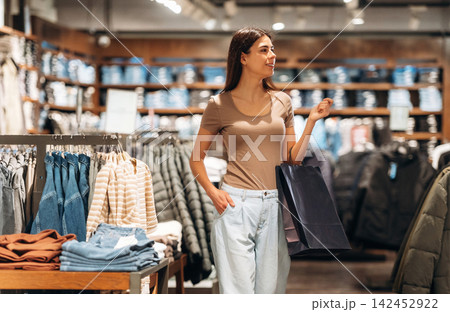 Young woman shopping in a clothing store, holding a shopping bag, surrounded by racks of apparel and folded garments in a well-lit retail environment Young woman shopping in a clothing store, holding a shopping bag, surrounded by racks of apparel and folded garments in a well-lit retail environment 142452922