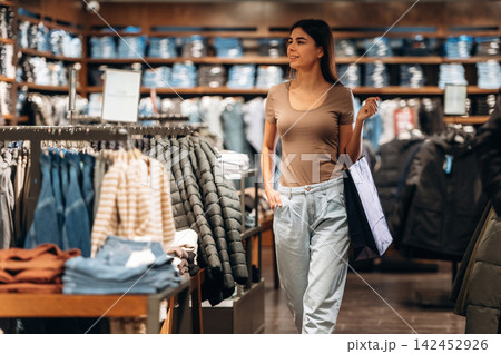 Female shopper carrying a shopping bag while browsing clothing racks inside a modern retail store with various garments displayed on shelves Female shopper carrying a shopping bag while browsing clothing racks inside a modern retail store with various garments displayed on shelves 142452926