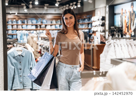 Young woman with long dark hair smiles while holding shopping bags inside a clothing store, racks of apparel and accessories visible in the background Young woman with long dark hair smiles while holding shopping bags inside a clothing store, racks of apparel and accessories visible in the background 142452970