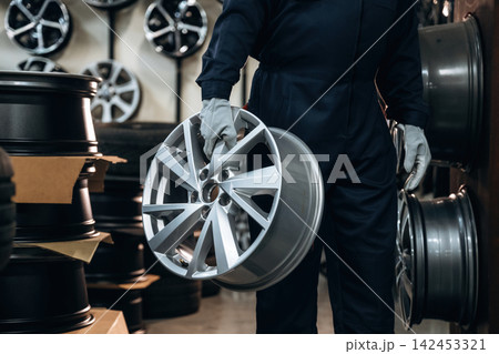 Male technician in dark coveralls holds a silver alloy wheel while surrounded by stacks of tires and additional wheels in a well-lit automotive workshop 142453321