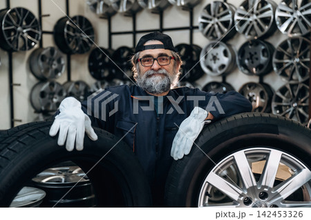 Senior male mechanic with glasses and cap holds two tires in a workshop, surrounded by various car rims displayed on the wall behind him Senior male mechanic with glasses and cap holds two tires in a workshop, surrounded by various car rims displayed on the wall behind him 142453326