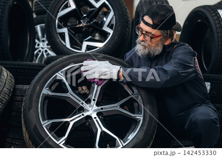 Senior male technician wearing glasses and gloves cleans a car wheel with a cloth in a tire shop surrounded by stacks of tires and wheels in the background Senior male technician wearing glasses and gloves cleans a car wheel with a cloth in a tire shop surrounded by stacks of tires and wheels in the background 142453330
