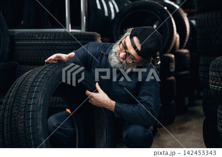 Elderly Caucasian male mechanic inspects a tire in a tire shop surrounded by stacks of tires, wearing a blue jumpsuit and a black cap, focused on the task at hand 142453343