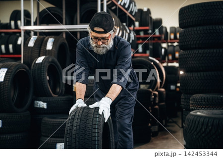 Senior male tire technician in dark coveralls lifts a tire in a storage area filled with stacked tires and shelves, showcasing a well-organized workshop environment 142453344