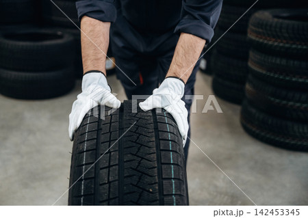 Male technician wearing gloves lifts a car tire in a workshop with stacks of tires visible in the background, showcasing automotive maintenance and repair environment Male technician wearing gloves lifts a car tire in a workshop with stacks of tires visible in the background, showcasing automotive maintenance and repair environment 142453345