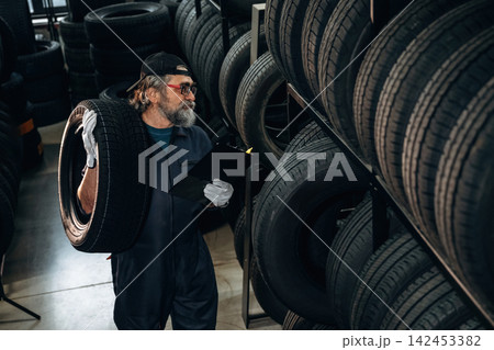Caucasian male tire technician wearing gloves inspects tire while holding clipboard in tire storage area with stacked tires in the background 142453382