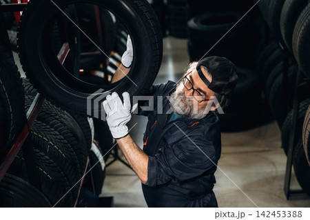 Senior male mechanic with glasses lifts a tire in a tire shop surrounded by stacked tires on shelves, wearing gloves and a cap, focused on his task Senior male mechanic with glasses lifts a tire in a tire shop surrounded by stacked tires on shelves, wearing gloves and a cap, focused on his task 142453388
