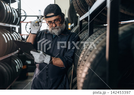 Senior male mechanic with glasses holding clipboard and pencil, standing among stacked tires in a well-organized automotive workshop with shelves in the background Senior male mechanic with glasses holding clipboard and pencil, standing among stacked tires in a well-organized automotive workshop with shelves in the background 142453397