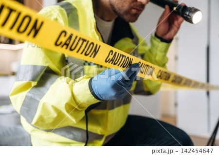 Forensic expert in yellow safety jacket examines caution tape with flashlight at a crime scene, highlighting the importance of evidence collection and investigation procedures Forensic expert in yellow safety jacket examines caution tape with flashlight at a crime scene, highlighting the importance of evidence collection and investigation procedures 142454647