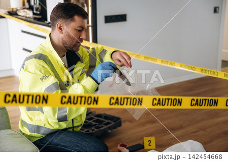Police detective examining evidence at a murder scene, wearing a reflective jacket, with caution tape and a body outline visible on the floor 142454668