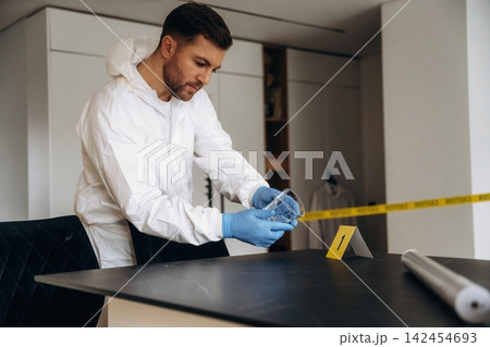 Forensic expert in white protective suit examining evidence at a crime scene, measuring with a tape measure on a black table with evidence markers visible Forensic expert in white protective suit examining evidence at a crime scene, measuring with a tape measure on a black table with evidence markers visible 142454693