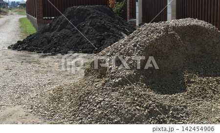Piles of construction materials on the side of a dirt road near a fence. In the foreground is a large mound of fine gravel and pebbles, and in the background is a pile of dark black soil. Piles of construction materials on the side of a dirt road near a fence. In the foreground is a large mound of fine gravel and pebbles, and in the background is a pile of dark black soil. 142454906
