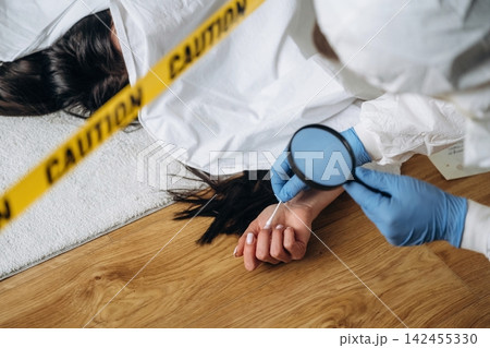 Forensic expert examining evidence at a murder scene with a magnifying glass, caution tape visible around the body on a wooden floor 142455330