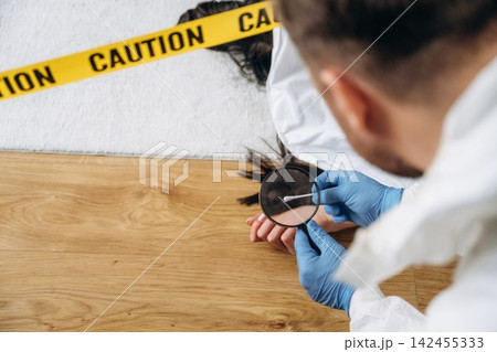 Forensic expert examining evidence at a murder scene, wearing gloves and protective clothing, with caution tape and a body visible in the background 142455333
