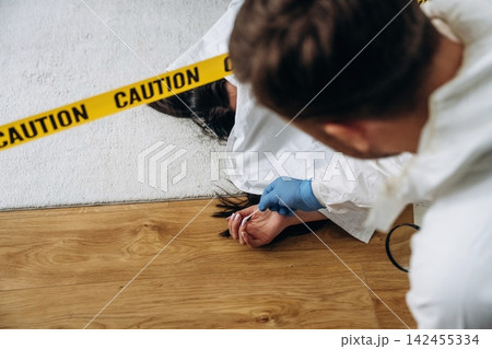 Forensic expert examining a crime scene with a body on the floor, surrounded by caution tape and evidence markers in a residential setting Forensic expert examining a crime scene with a body on the floor, surrounded by caution tape and evidence markers in a residential setting 142455334