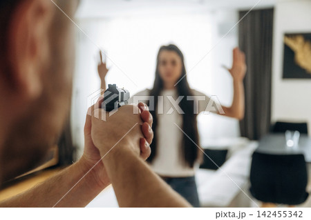 Man holding a handgun aimed at a woman with raised hands in a domestic setting, with a sofa and table visible in the background Man holding a handgun aimed at a woman with raised hands in a domestic setting, with a sofa and table visible in the background 142455342