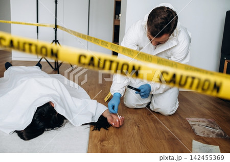 Forensic expert in white suit examines evidence at a murder scene with caution tape surrounding a body on the floor in a residential setting 142455369
