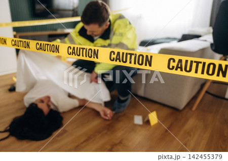 Forensic expert in yellow jacket examines body at a murder scene, with caution tape and evidence markers visible on the wooden floor in a residential setting Forensic expert in yellow jacket examines body at a murder scene, with caution tape and evidence markers visible on the wooden floor in a residential setting 142455379