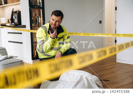 Police detective in reflective jacket kneeling near a covered body at a murder scene, surrounded by caution tape in a modern apartment interior Police detective in reflective jacket kneeling near a covered body at a murder scene, surrounded by caution tape in a modern apartment interior 142455386