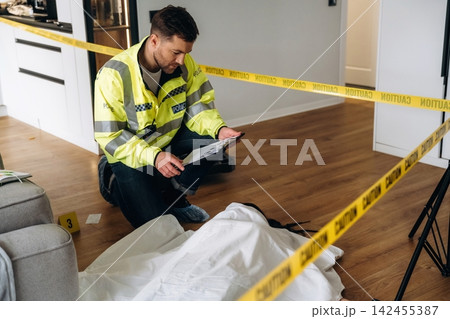Police detective in reflective jacket examines evidence at a murder scene marked by caution tape, with a covered body on the floor and a modern interior setting 142455387