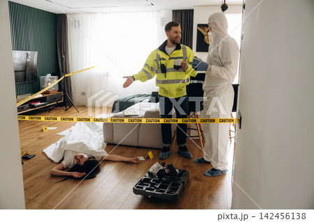 Forensic expert in white suit examines evidence at a murder scene, while police detective in reflective vest discusses findings in a modern apartment interior Forensic expert in white suit examines evidence at a murder scene, while police detective in reflective vest discusses findings in a modern apartment interior 142456138