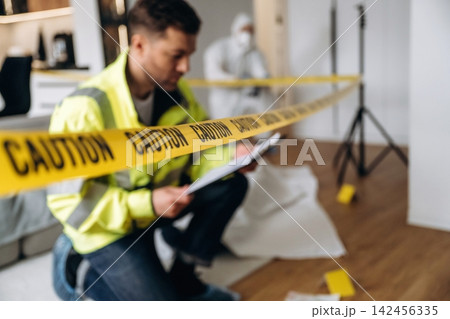 Forensic expert in yellow safety jacket kneeling near caution tape while examining evidence at a crime scene with a figure in protective suit in the background Forensic expert in yellow safety jacket kneeling near caution tape while examining evidence at a crime scene with a figure in protective suit in the background 142456335
