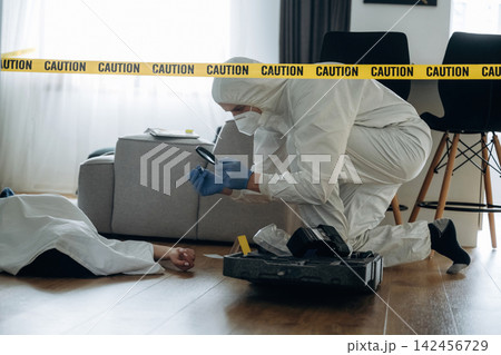 Forensic expert in protective suit examining evidence at a murder scene, with caution tape and a body on the floor in a residential setting 142456729