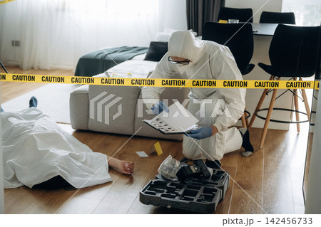 Forensic expert in protective suit examines evidence at a murder scene with caution tape, a body covered in white sheet, and a crime scene investigation kit nearby Forensic expert in protective suit examines evidence at a murder scene with caution tape, a body covered in white sheet, and a crime scene investigation kit nearby 142456733