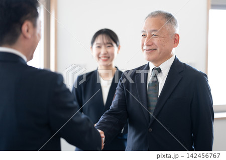 オフィスで笑顔で握手を交わすスーツ姿のシニアビジネスパーソンと見守る女性社員 オフィスで笑顔で握手を交わすスーツ姿のシニアビジネスパーソンと見守る女性社員 142456767
