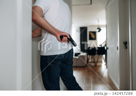 Male figure in white t-shirt holding a handgun behind his back while standing in a modern interior hallway with wooden flooring and contemporary furniture visible 142457289