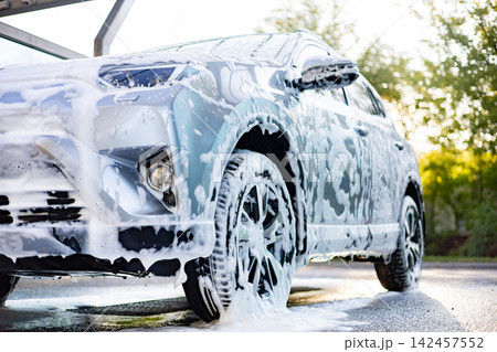 A silver SUV is covered in thick white foam during a thorough car wash process outdoors A silver SUV is covered in thick white foam during a thorough car wash process outdoors 142457552