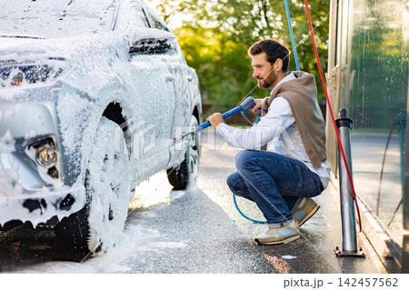 A man is kneeling down and using a pressure washer to clean his car covered in foam A man is kneeling down and using a pressure washer to clean his car covered in foam 142457562