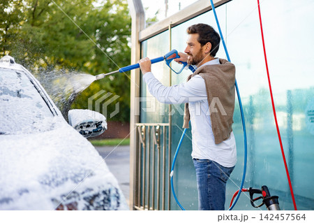A smiling Caucasian man uses a high-pressure washer to clean his car covered in foam at an outdoor self-service car wash station A smiling Caucasian man uses a high-pressure washer to clean his car covered in foam at an outdoor self-service car wash station 142457564