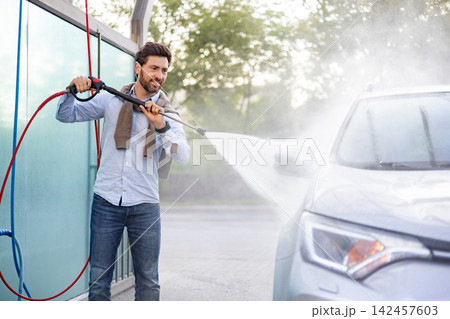 A smiling caucasian man in jeans and a blue shirt uses a high-pressure water spray to clean his car at an outdoor wash station A smiling caucasian man in jeans and a blue shirt uses a high-pressure water spray to clean his car at an outdoor wash station 142457603