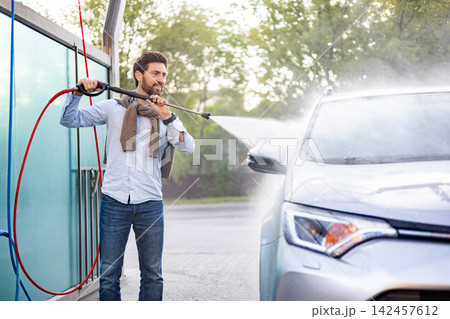 A man is using a high-pressure water jet to clean his car at an outdoor self-service car wash station 142457612