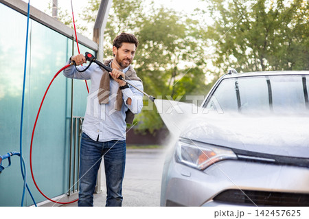 A man is actively cleaning his car using a high-pressure water spray at an outdoor self-service car wash station A man is actively cleaning his car using a high-pressure water spray at an outdoor self-service car wash station 142457625