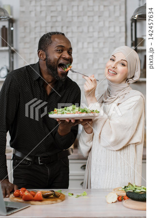 A African man and a Muslim woman share a moment of joy while preparing and eating a fresh salad in a bright kitchen setting 142457634