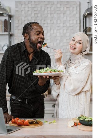 A man and woman of diverse backgrounds share a moment of joy while preparing and tasting a fresh salad in a bright kitchen setting 142457635