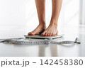 Barefoot individual standing on a digital scale with a measuring tape lying on the floor, bright indoor setting with natural light illuminating the scene 142458380