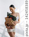 Slim woman in white sportswear holding a paper bag filled with fresh vegetables and fruits, standing in a bright indoor space with natural light streaming through windows 142458389