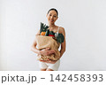 Young woman holding a large paper bag filled with fresh vegetables and fruits, wearing a white tank top and shorts, standing against a plain white background 142458393