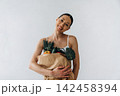 Slim woman with dark hair holds a paper bag filled with fresh vegetables, smiling against a light gray background in a minimalist indoor setting 142458394