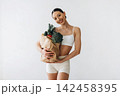 Slim female model in white sportswear holds a paper bag filled with fresh vegetables, standing against a plain white background in a bright indoor setting 142458395