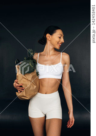 Female model in white sports bra and shorts holds a paper bag filled with fresh vegetables, showcasing a healthy lifestyle against a dark background 142458401