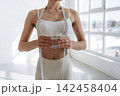 Slim female figure holding a glass of water while measuring her waist with a tape measure in a bright, minimalistic indoor space with large windows 142458404
