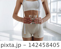 Slim female figure in white sportswear holds a glass of water while measuring her waist with a tape measure in a bright, minimalistic indoor setting 142458405