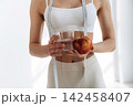 Slim female figure holding a glass of water and a red apple, measuring tape draped around her waist, bright indoor setting with soft natural light 142458407